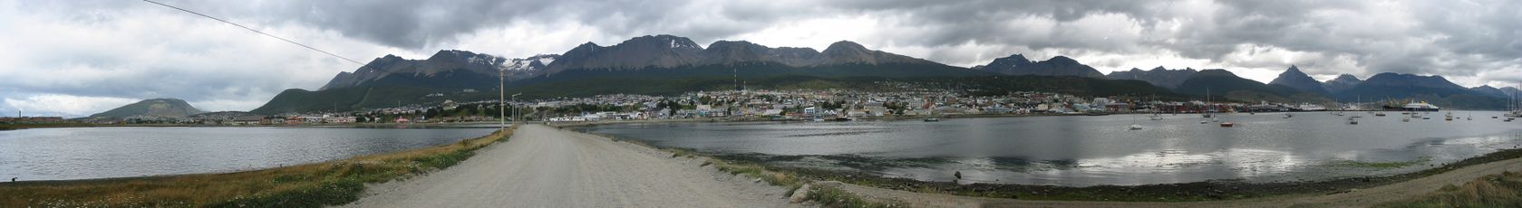 A small city across a gray waterway under lowering gray clouds. A road leads to the city across a causeway. Mountains with snow and a low treeline form the backdrop. A few boats are in the water.